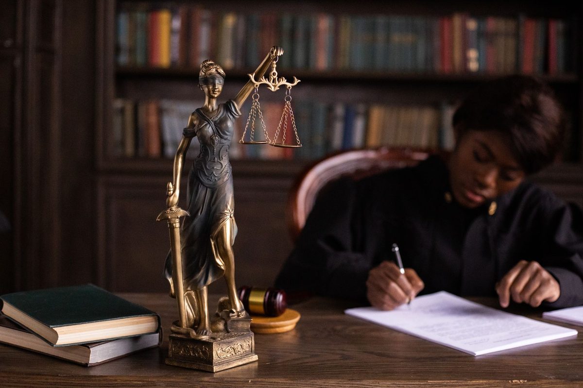 Estátua da Justiça em primeiro plano diante de uma magistrada em biblioteca jurídica