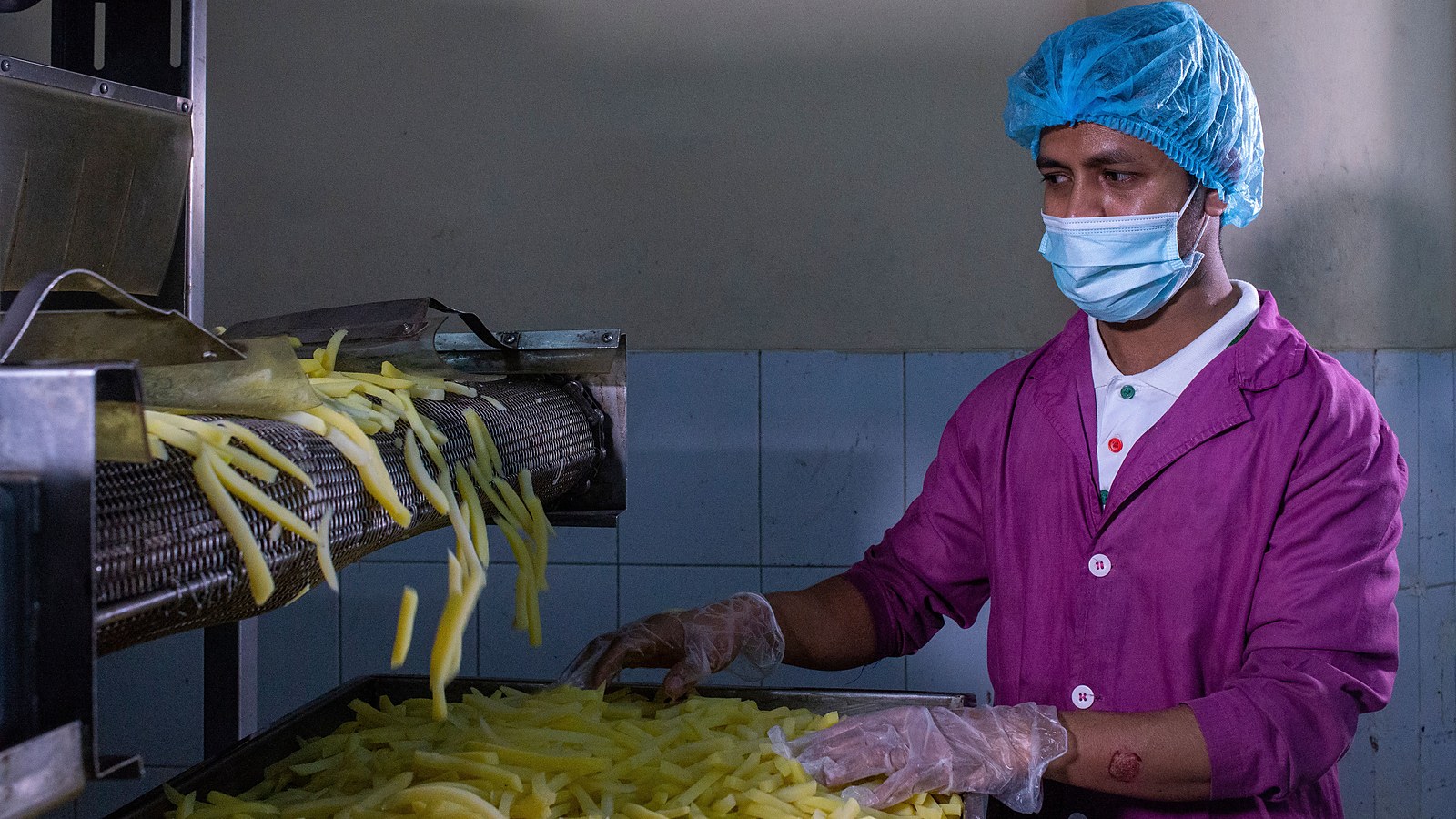 Operário de touca, máscara e luvas descartáveis acompanha a esteira de uma linha de produção de alimentos