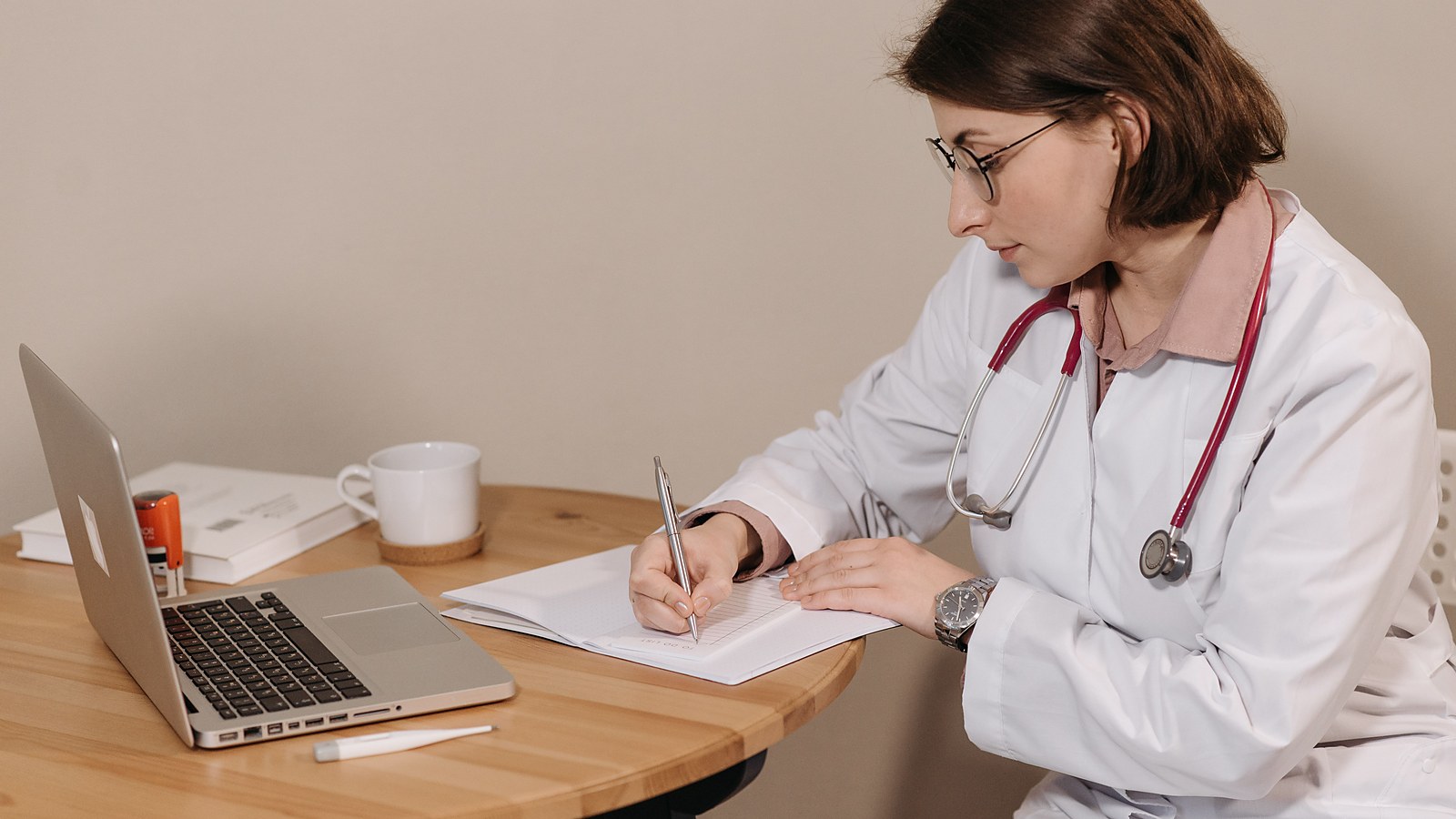 Médica de jaleco branco preenche formulários à mesa, diante do computador, sem paciente na sala