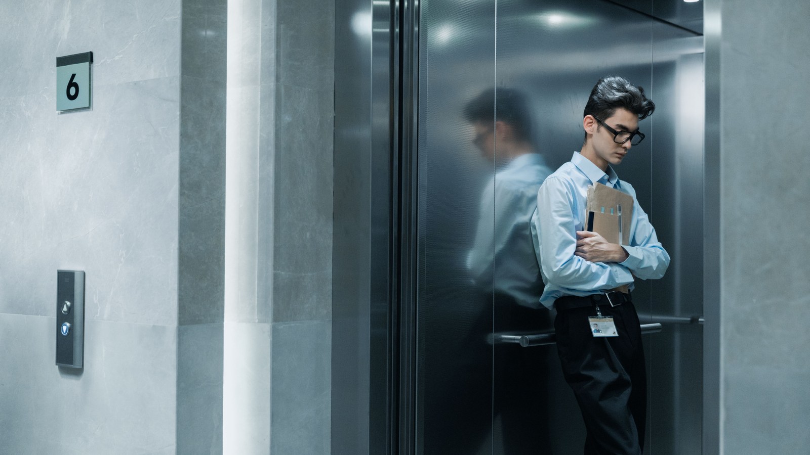 Homem de camisa e gravata, de cabeça baixa, segura envelopes de papel diante da porta do elevador, cena que ilustra a demissão humilhante discutida no artigo.