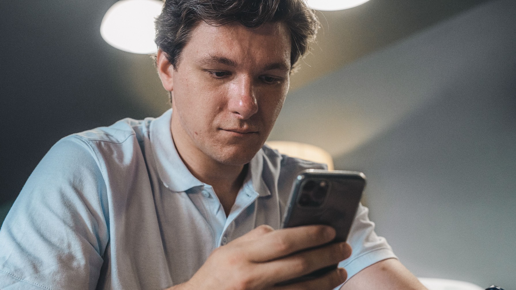 Homem adulto sentado em casa, de camisa clara, segura um telefone celular com uma das mãos e mantém o olhar baixo, voltado para o aparelho.