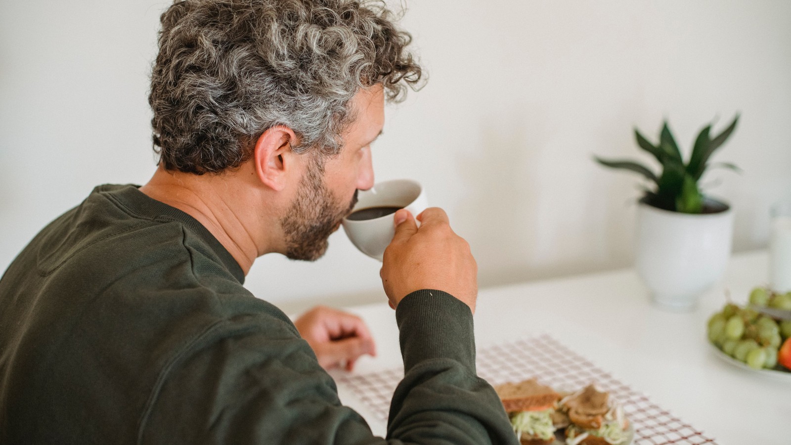 Homem adulto de barba grisalha, sentado sozinho à mesa de uma cozinha clara, bebe de uma xícara branca ao lado de um prato de sanduíche.
